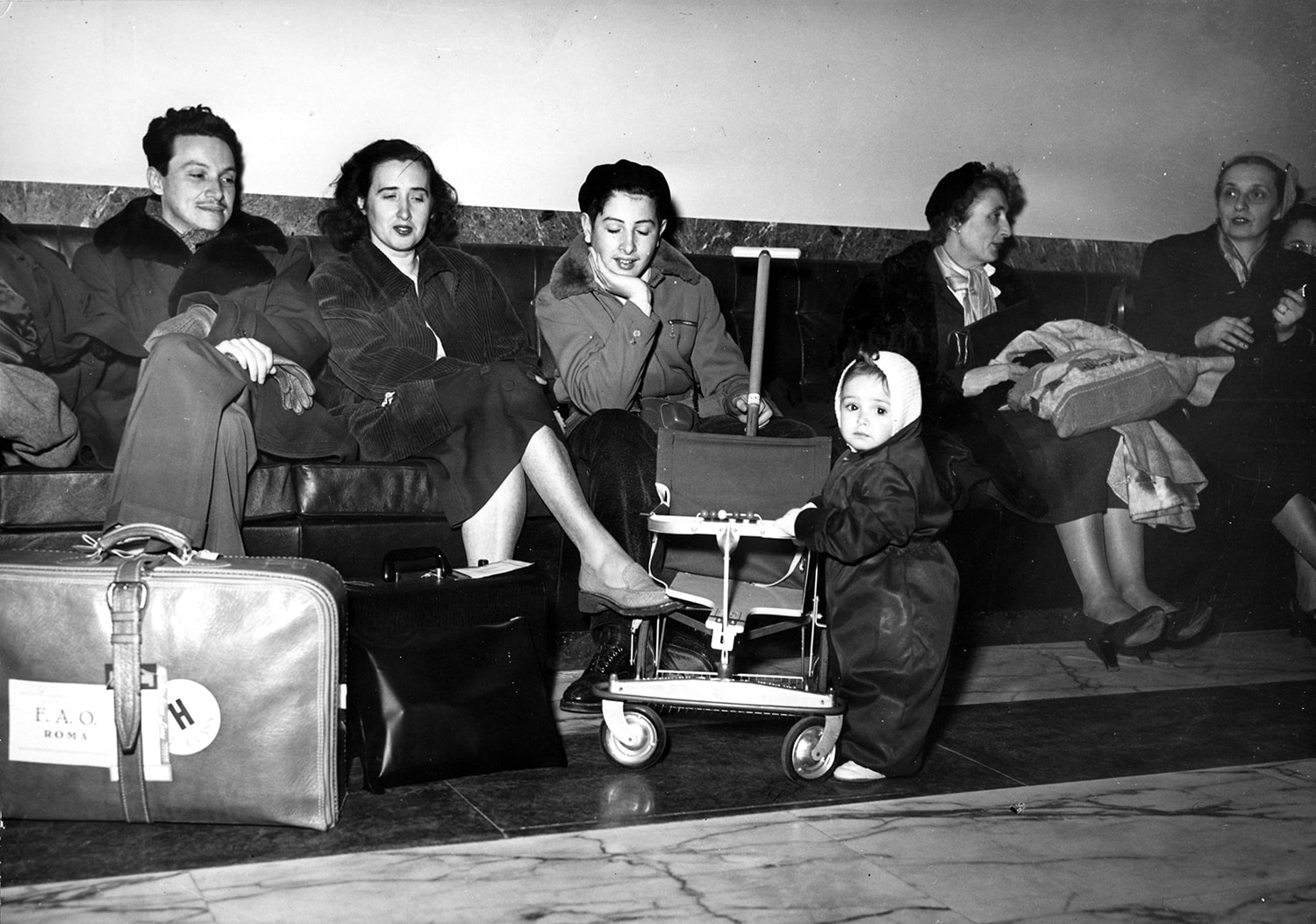 Four women, one man and a young child await transfer among pieces of luggage