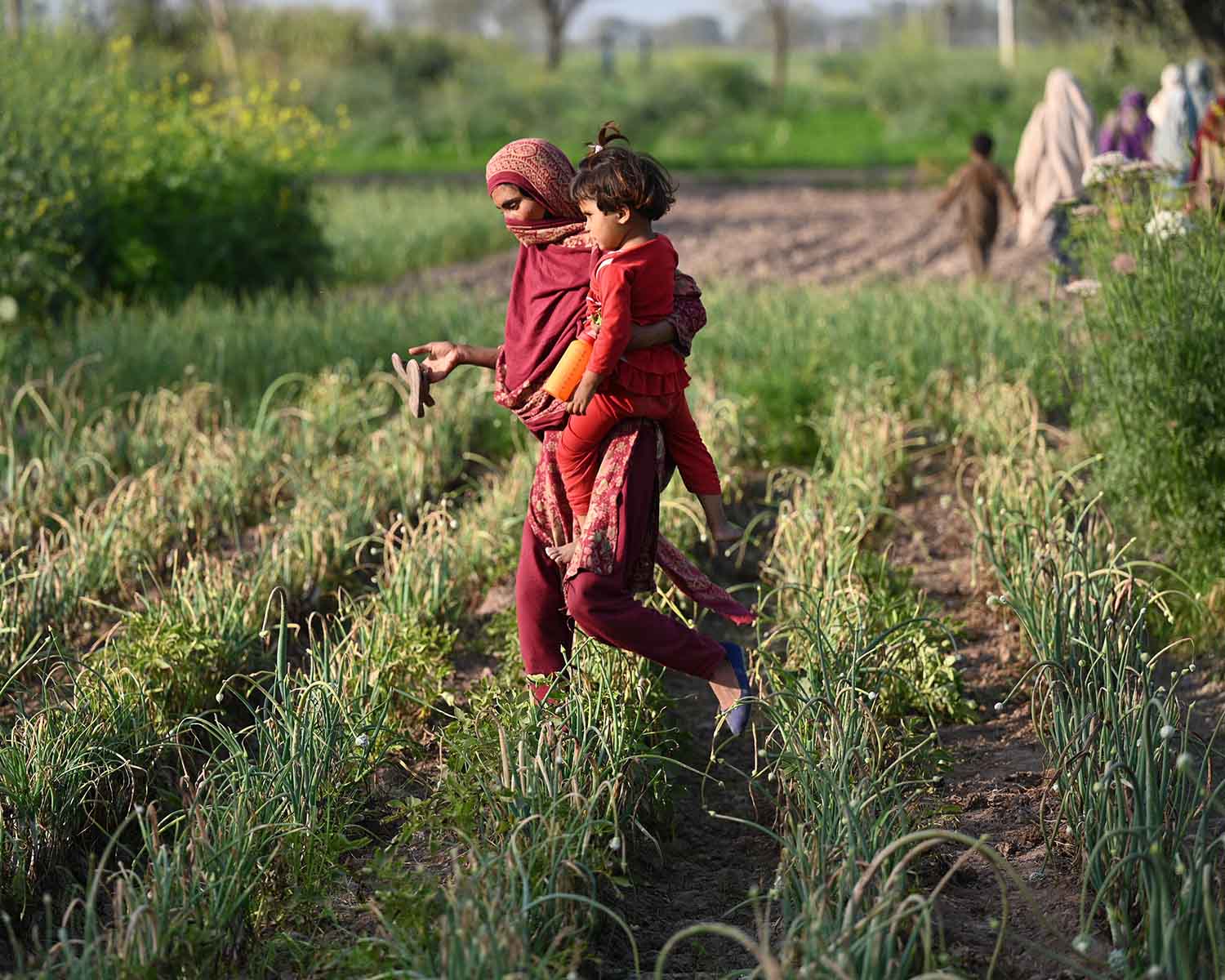 Pakistan – A woman holds her daughter as she arrives to work at a vegetable field.