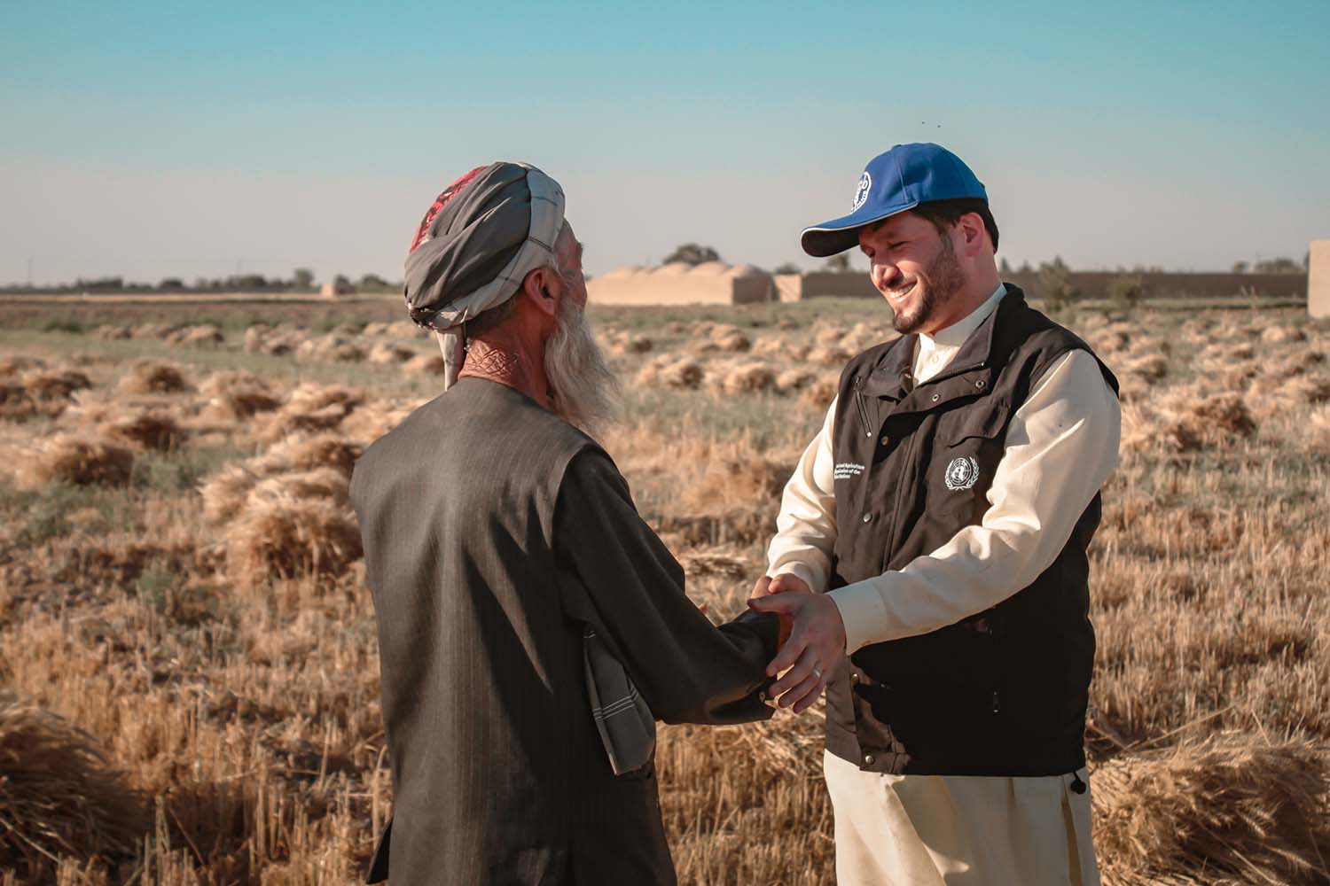 An FAO engineer in a cap and tunic shakes hands with an Afghan farmer in traditional dress.